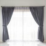 Rideaux gris élégamment tirés de chaque côté d'une baie vitrée, avec voilage blanc translucide au centre, dans un salon moderne et lumineux.