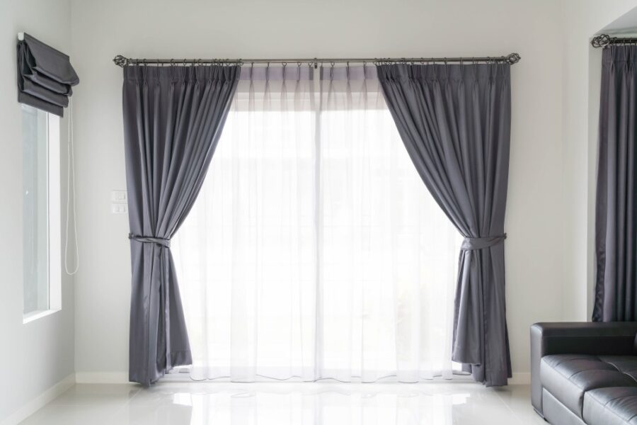 Habillez vos ouvertures pour allier isolation et décoration Rideaux gris élégamment tirés de chaque côté d'une baie vitrée, avec voilage blanc translucide au centre, dans un salon moderne et lumineux.
