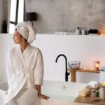 Femme en peignoir blanc assise sur une baignoire dans une salle de bain spa moderne avec bougies, produits de soin et lumière tamisée