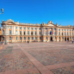 Vue de la place du Capitole à Toulouse, avec la façade de l'hôtel de ville, son architecture en briques roses et sa grande esplanade pavée.