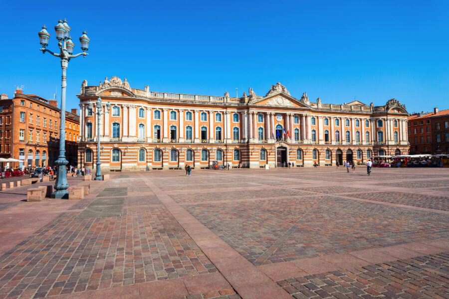 Vue de la place du Capitole à Toulouse, avec la façade de l'hôtel de ville, son architecture en briques roses et sa grande esplanade pavée.